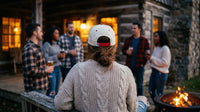 Group of people gathered around a fire pit at dusk, with one person wearing a white cap with red accents.